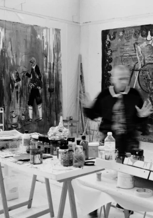 Black and white image inside artist studio with a blurred man, art supplies on a table, and two large paintings hanging on the wall.