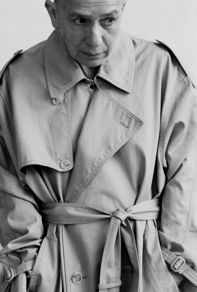 Monochrome portrait of a mature adult with a bald head wearing a belted trench coat, set against a plain white background in a studio.