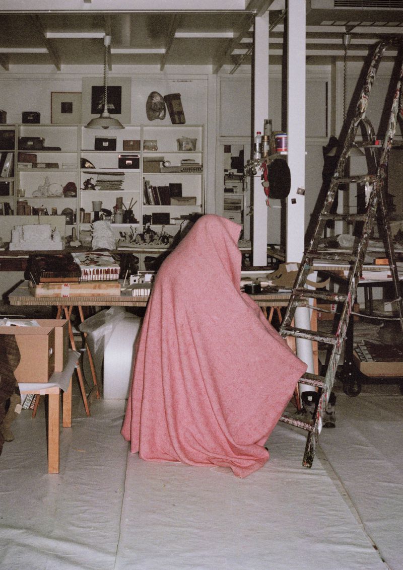 Person draped in a pink sheet stands in a cluttered studio with ladders, shelves, and various art supplies, creating an enigmatic presence.
