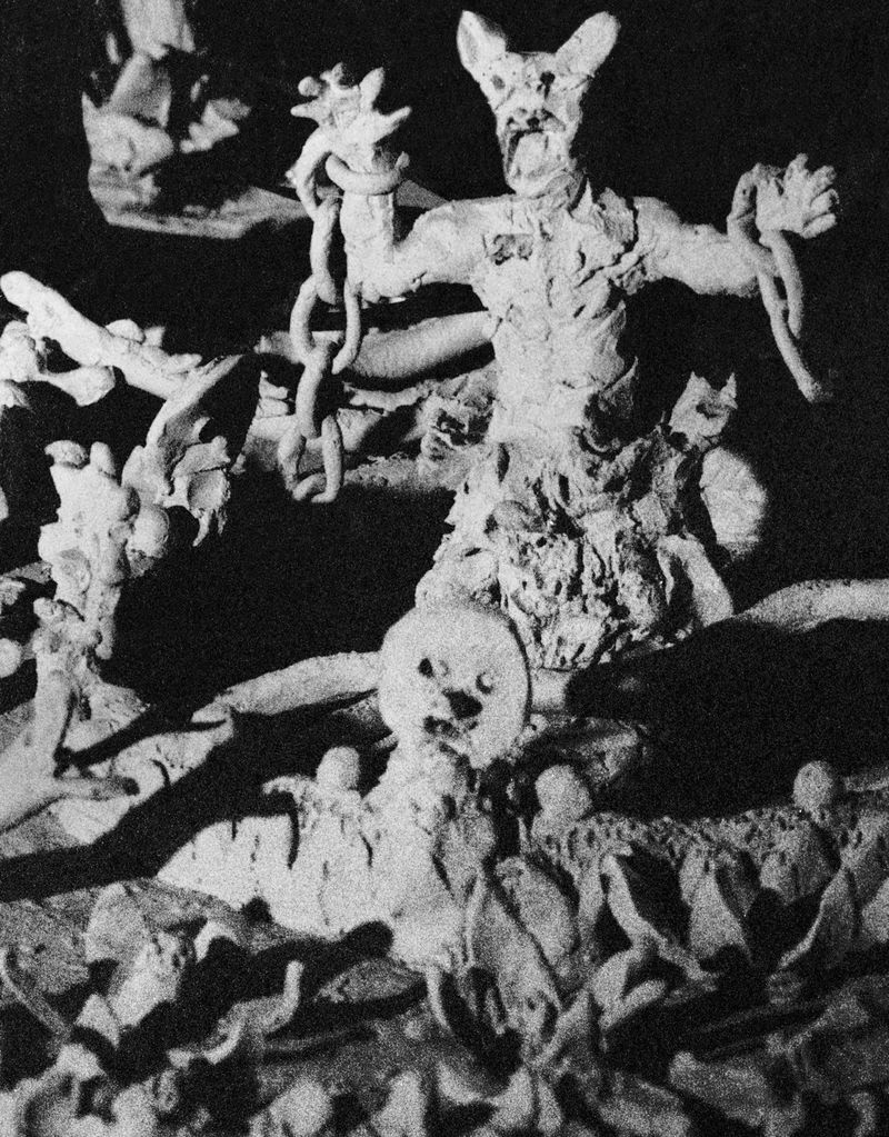 Monochrome sculpture of a horned figure with chains, standing on a base of skulls and figures, rendered in a grainy, dark style.