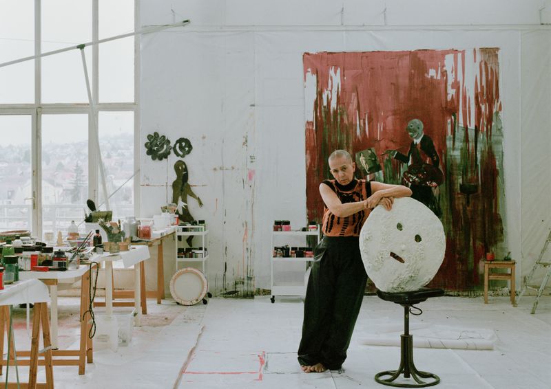 Artist in studio stands with sculpture. Large abstract painting hangs in background. Table with paint supplies and bright window light.