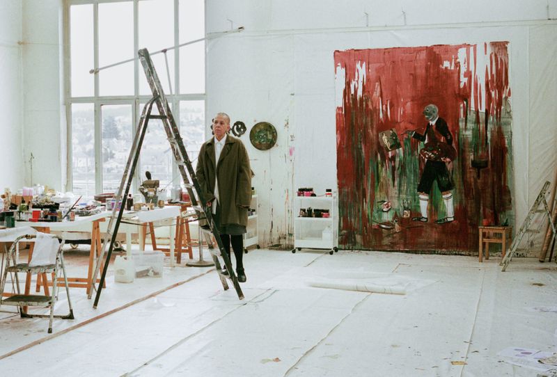 Adult male stands on a ladder in an artist's studio with large paintings and art supplies, under natural light from a large window.