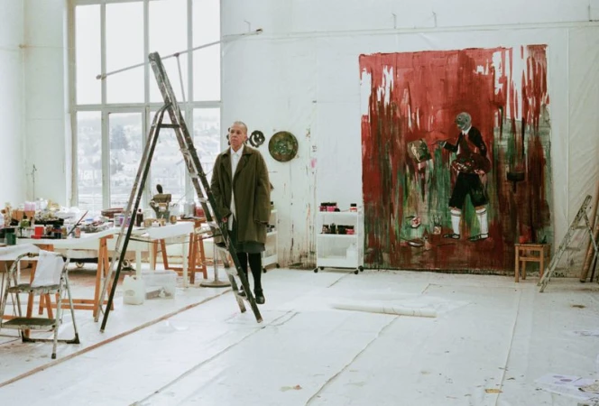 Adult male stands on a ladder in an artist's studio with large paintings and art supplies, under natural light from a large window.