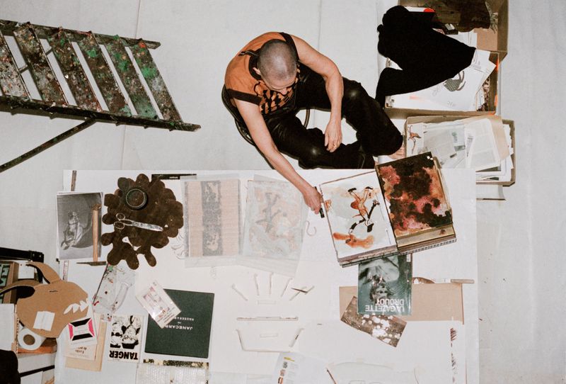 Overhead view of a person in a vest and leather pants standing over a table covered with art materials in a bright, white room.