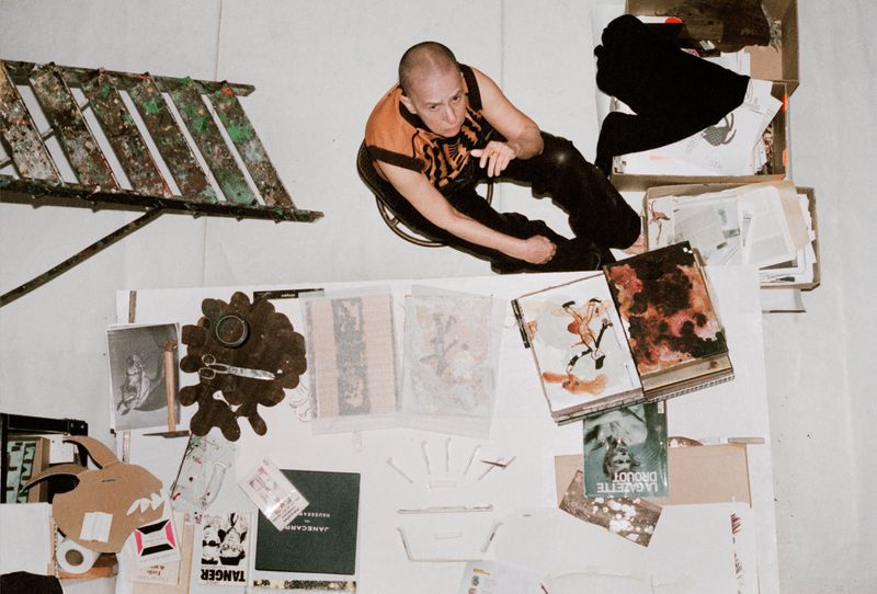 Overhead shot of a bald person in black and orange clothing sitting near a ladder and collection of books and papers on a white surface.