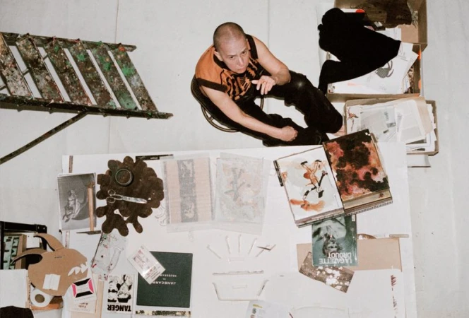 Overhead shot of a bald person in black and orange clothing sitting near a ladder and collection of books and papers on a white surface.