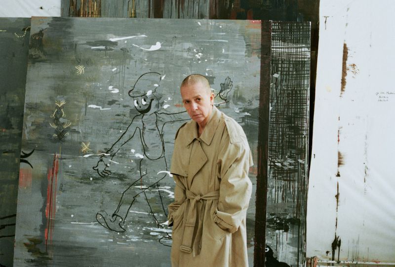 Adult female poses in front of a large abstract painting of a cartoon figure, wearing a tan trench coat in a white art studio.