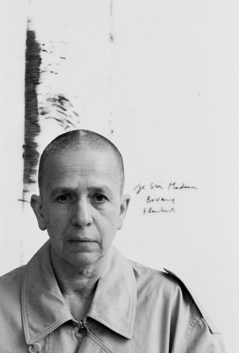 Black and white portrait of an adult person with short hair wearing a jacket, standing indoors in front of a white wall with text.