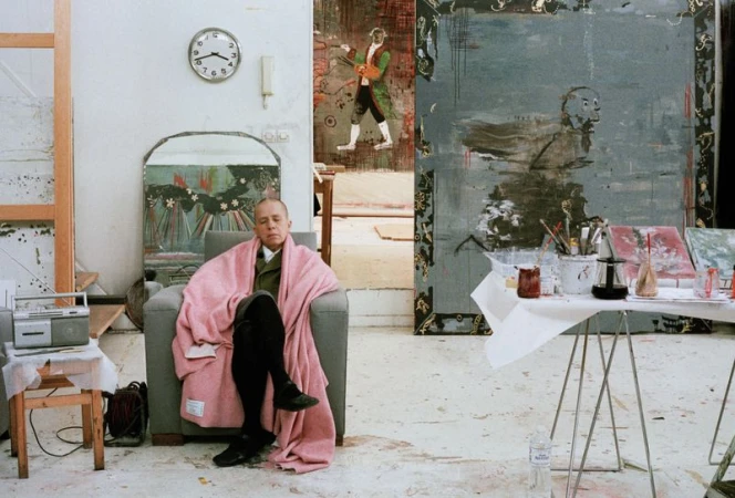Indoor portrait of an adult male artist sitting in a gray armchair with a pink blanket in an artist studio filled with artwork.