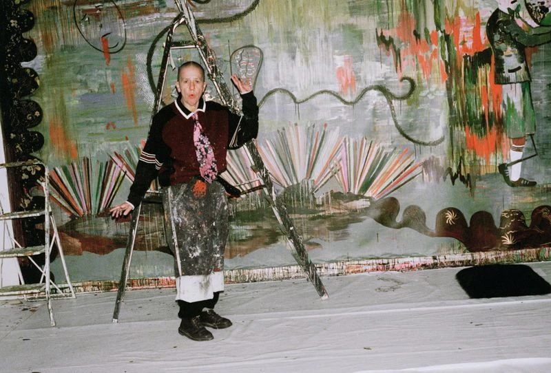 An adult female artist stands in front of her large-scale colorful abstract painting on a ladder in a studio or art space.