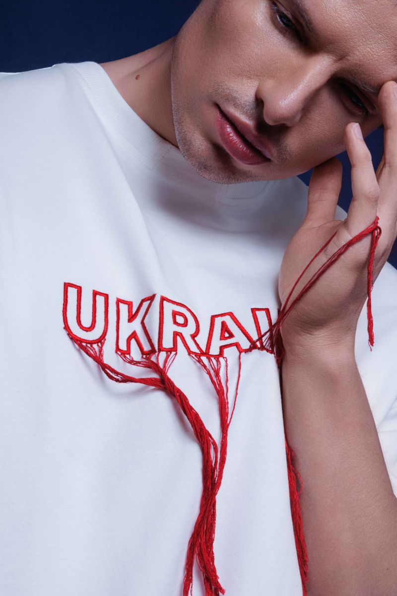 Close-up of a pensive adult male in a white t-shirt with embroidered 'Ukraine' and red threads, set against a dark blue background.