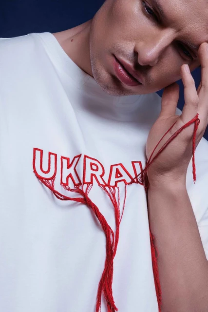 Close-up of a pensive adult male in a white t-shirt with embroidered 'Ukraine' and red threads, set against a dark blue background.