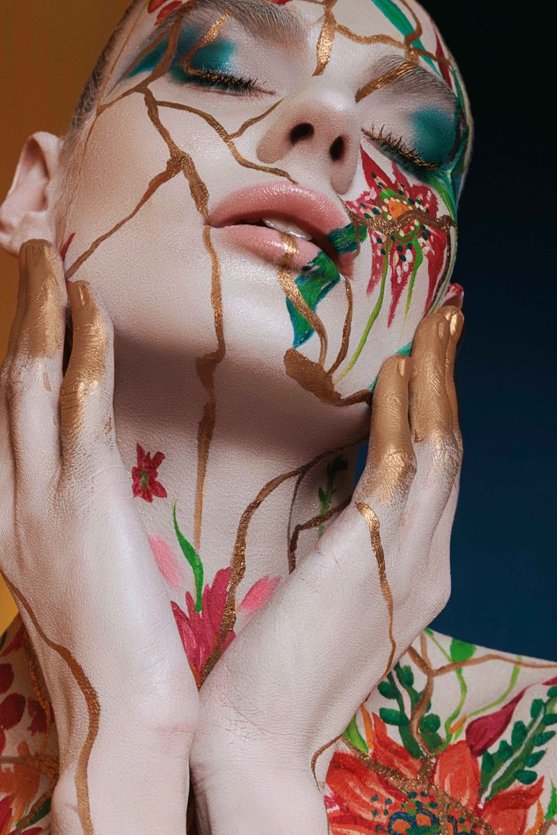 Artistic closeup portrait of an adult woman with dramatic floral body paint and gold line details on her face and hands on a gradient background.