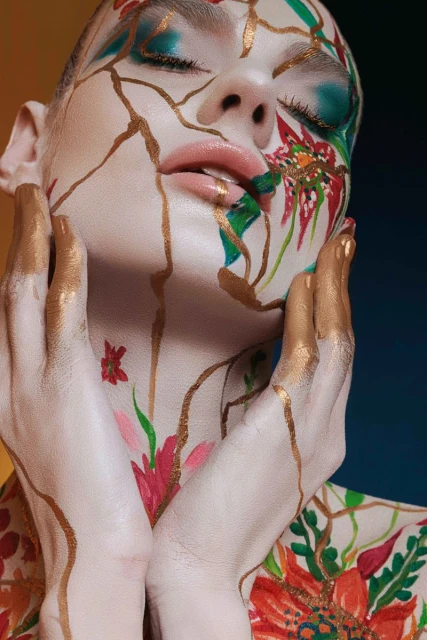 Artistic closeup portrait of an adult woman with dramatic floral body paint and gold line details on her face and hands on a gradient background.