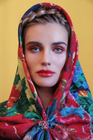 Close-up portrait of a woman wearing a vibrant floral headscarf with red, blue, and yellow patterns against a yellow background.