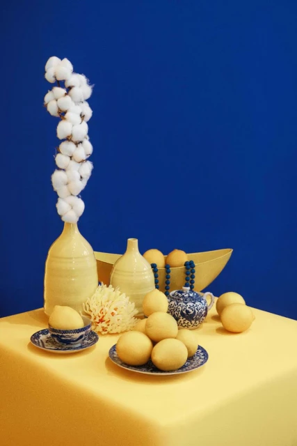 Still life with yellow vases, decorative bowl, lemons, and beaded decoration on yellow fabric against a blue backdrop.