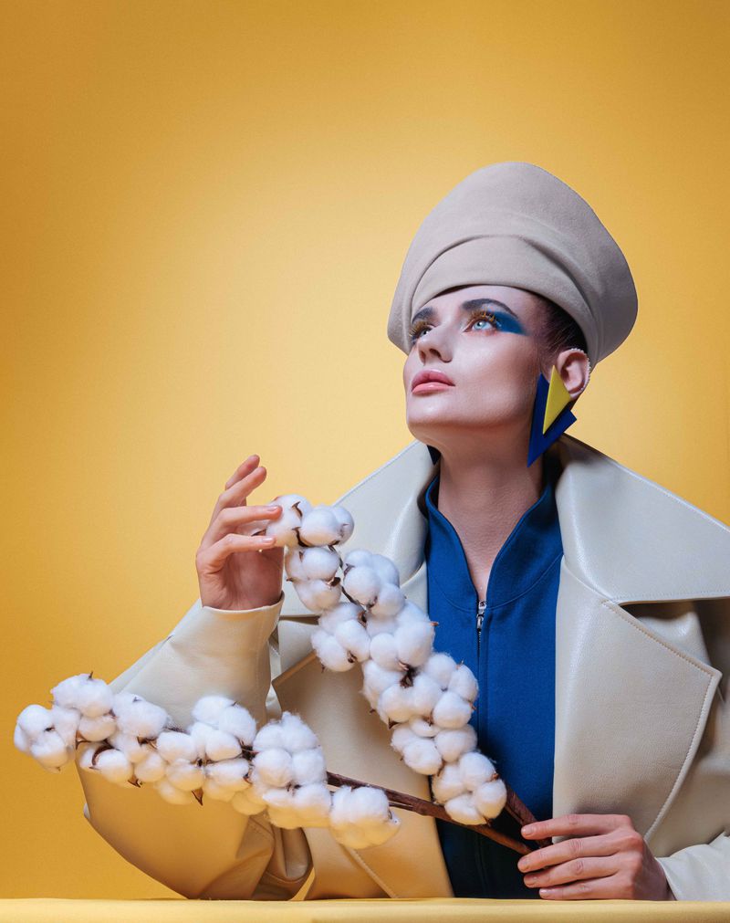 Fashionable woman in a beret and coat poses with cotton branches against a yellow backdrop, featuring blue and yellow geometric earrings.
