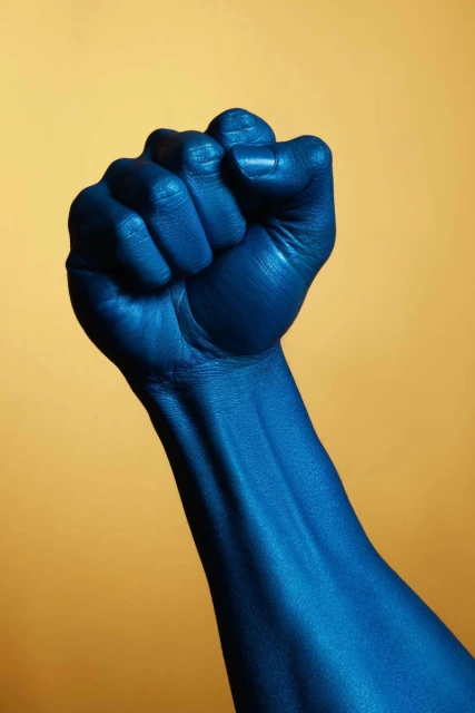 Close-up shot of an arm and hand painted in metallic blue, raised in a fist against a solid gold background, studio shot.