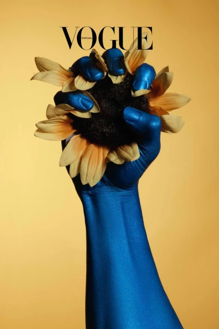 Editorial fashion cover featuring a blue-painted arm holding a sunflower against a yellow backdrop, featuring the Vogue magazine masthead.