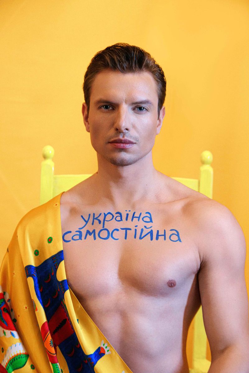 Portrait of a blond male model against a yellow background, wearing a printed scarf and body paint, looking directly at the camera.