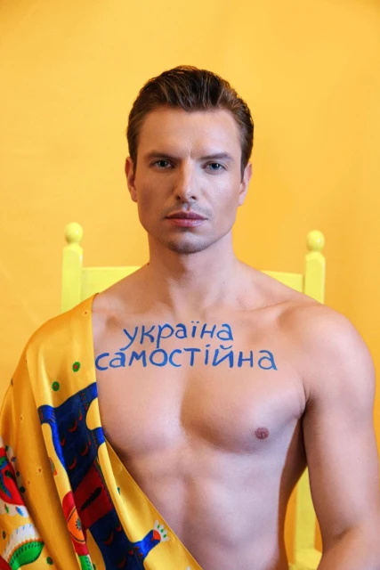 Portrait of a blond male model against a yellow background, wearing a printed scarf and body paint, looking directly at the camera.