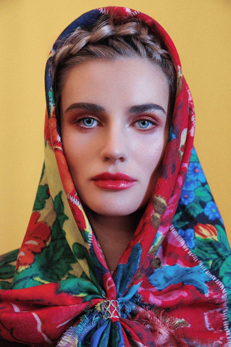 Close-up portrait of a woman wearing a vibrant floral headscarf with red, blue, and yellow patterns against a yellow background.