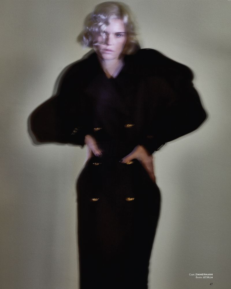 A woman in a long, black, double-breasted dress or coat poses with her hands on her hips in a blurred fashion studio shot.