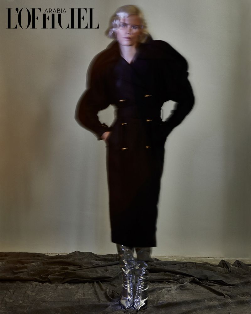 A model poses in a full-length shot wearing a black coat and metallic boots, under studio lighting, for a fashion editorial, cropped to the logo.