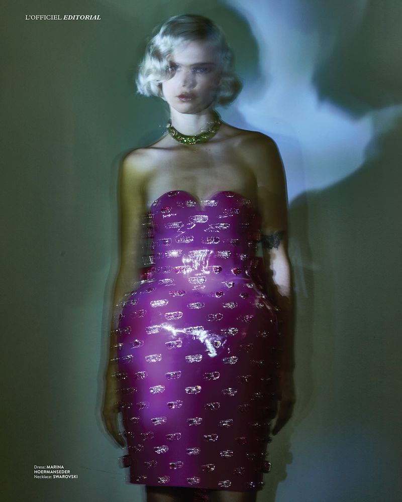 Blonde woman in a purple strapless dress and necklace with silver embellishments, posing in a soft, ethereal light for a fashion editorial.
