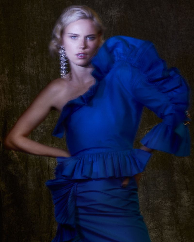 Woman in a blue one-shoulder dress with ruffle detail, posing against a blurred brown backdrop with long chandelier earrings.