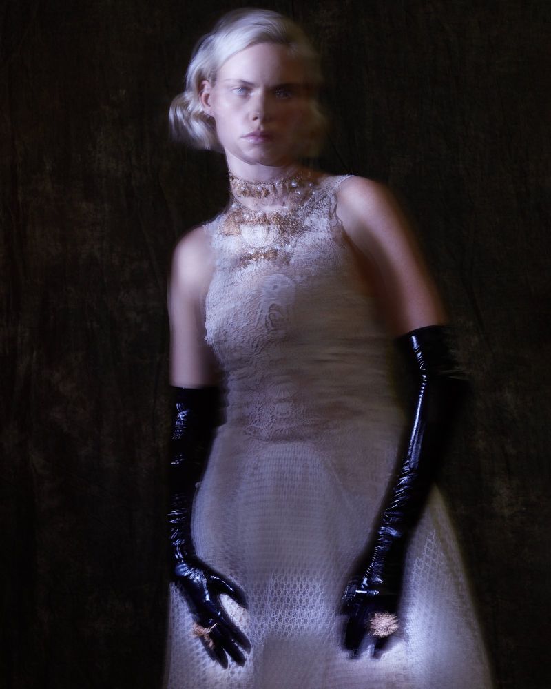 Elegant woman in a white lace dress, black gloves, and ornate jewelry posing in a photo studio with dark background, fashionable and sophisticated.