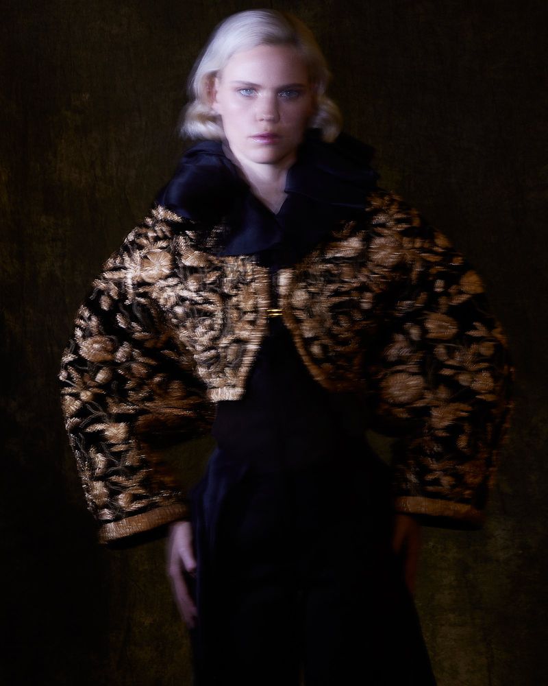 A woman with platinum blonde hair wears a black and gold embroidered jacket with a high collar against a dark backdrop.