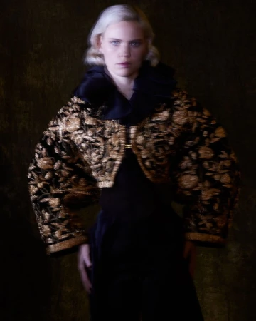 A woman with platinum blonde hair wears a black and gold embroidered jacket with a high collar against a dark backdrop.