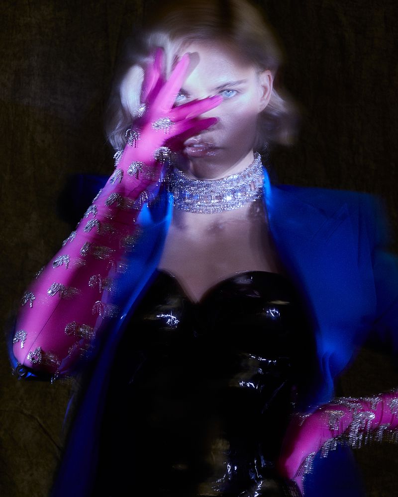 Model wearing vibrant pink embellished gloves over face with blue blazer and diamond choker necklace in editorial portrait.