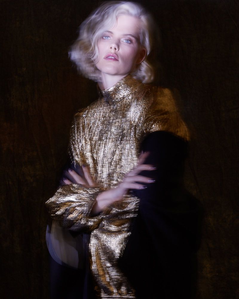 Portrait of an adult female with platinum blonde wavy hair, wearing a gold lame top with her arms crossed against a dark background.