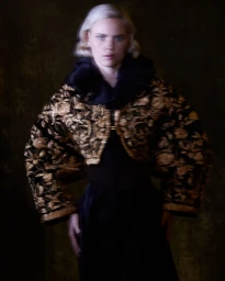Model with blonde hair wearing an embroidered gold and black jacket against a dark mottled background
