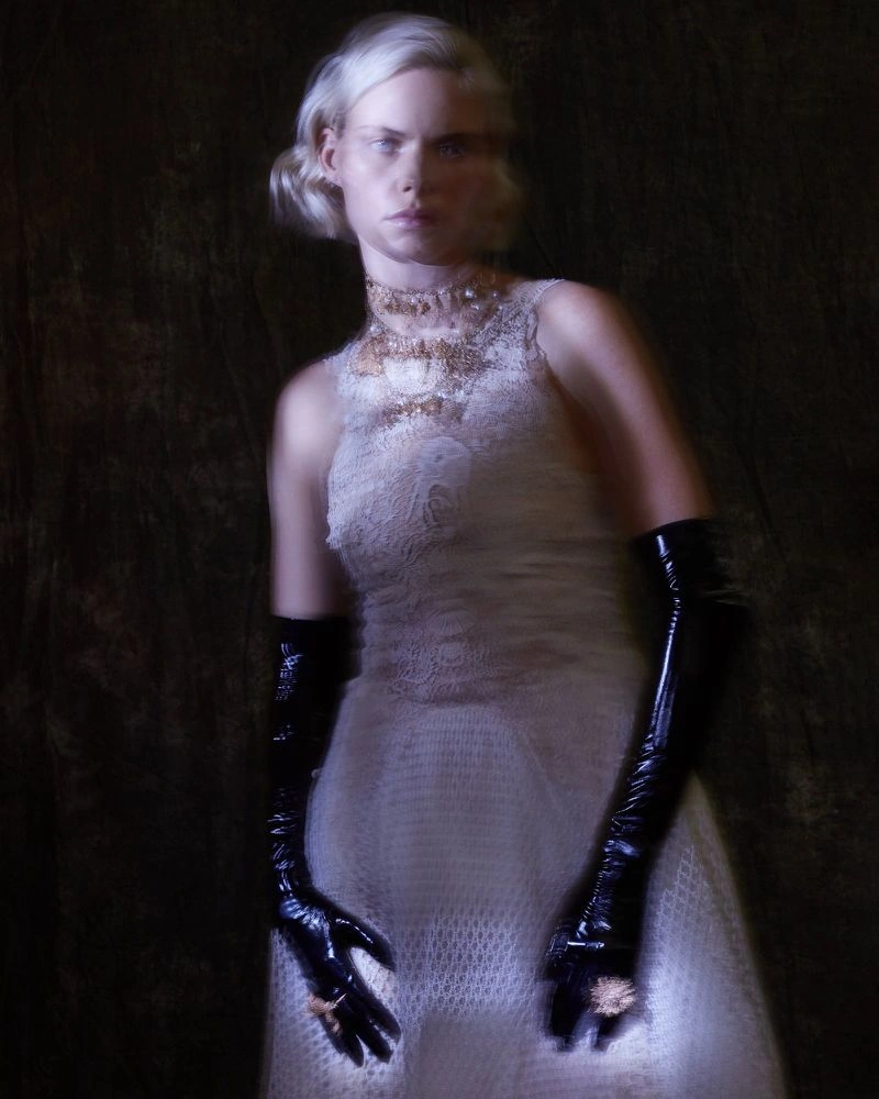 Model wearing a white lace dress and long black gloves posing against a dark textured background in low-key lighting.