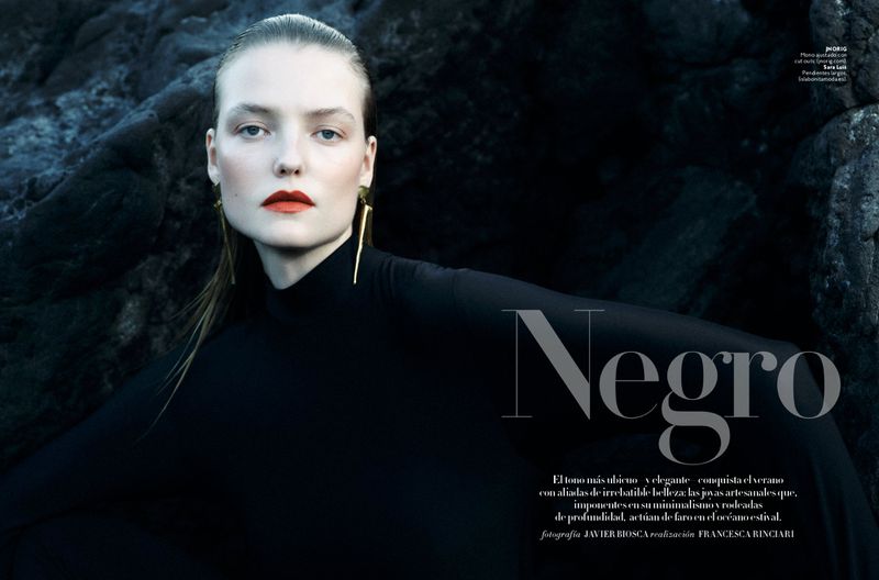 A woman with striking red lips and long gold earrings poses in front of a dark, rocky backdrop, wearing a sleek black top.