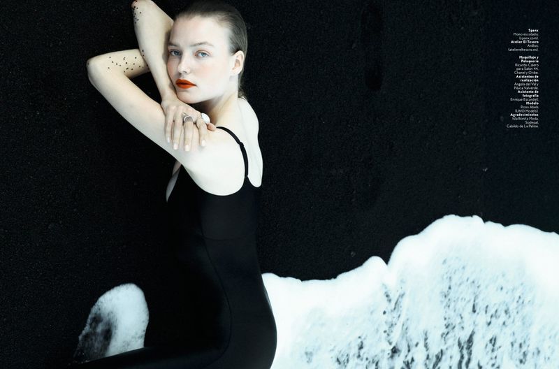 Woman in black swimsuit lying on black sand, posing with arms up. Dark background, light skin, red lips, and ocean foam visible.
