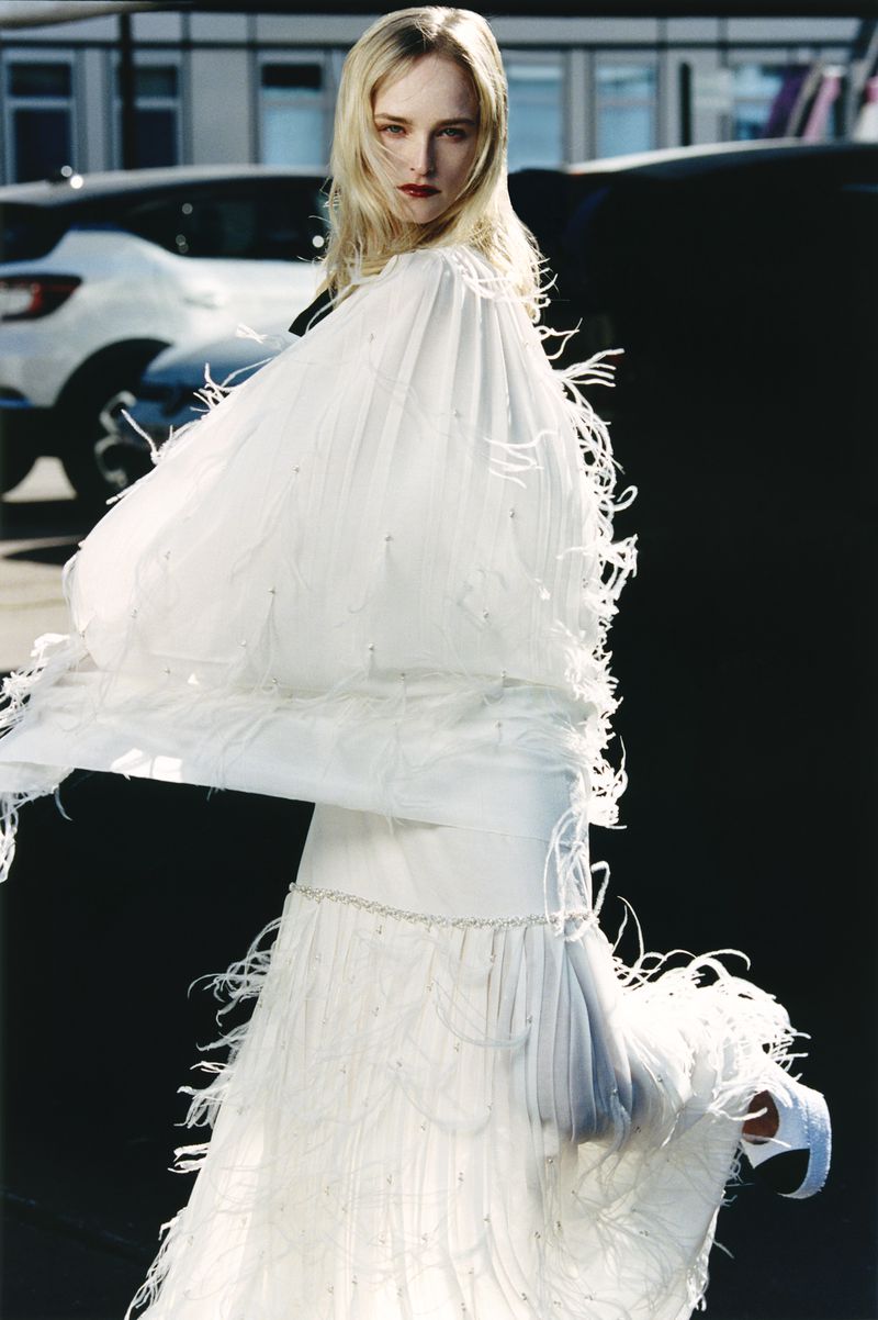 Fashion editorial shot of a blonde woman in a white feathered gown standing outdoors on a city street, cars and buildings behind her.