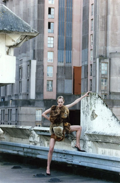 Fashion model in fur-trimmed dress poses on a rooftop with high heels and tall buildings in the background during daytime.