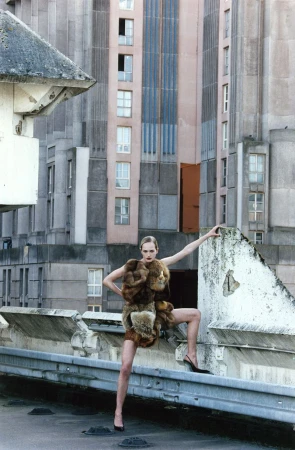 Fashion model in fur-trimmed dress poses on a rooftop with high heels and tall buildings in the background during daytime.