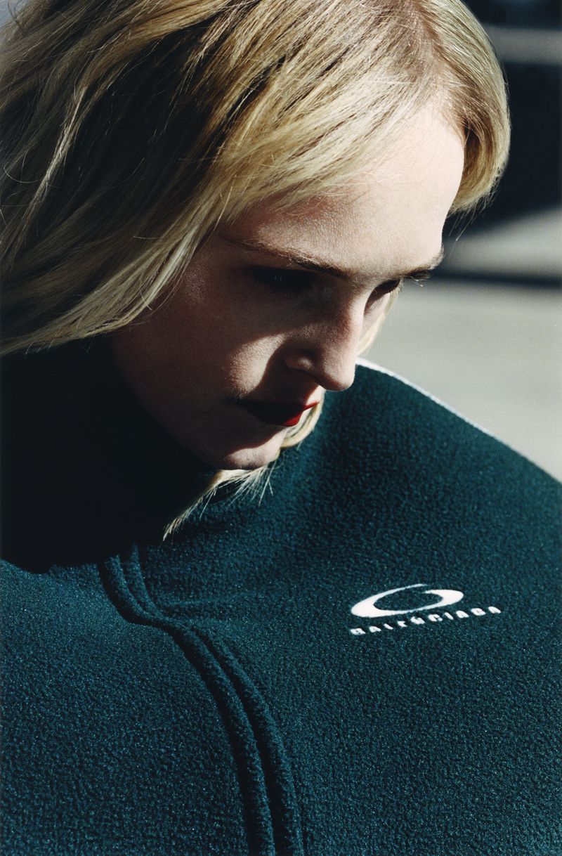 Close-up studio shot of a fair-skinned blonde woman wearing a dark green Balenciaga zip-up fleece with dark red lipstick.