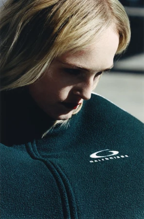 Close-up studio shot of a fair-skinned blonde woman wearing a dark green Balenciaga zip-up fleece with dark red lipstick.