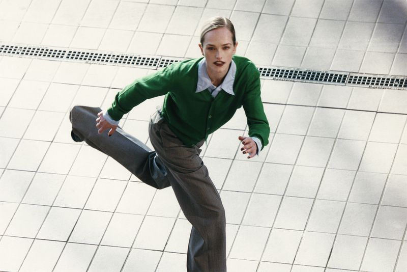 Woman in green cardigan and grey pants rides a skateboard across a tiled floor, shot from a high angle, bright natural light.