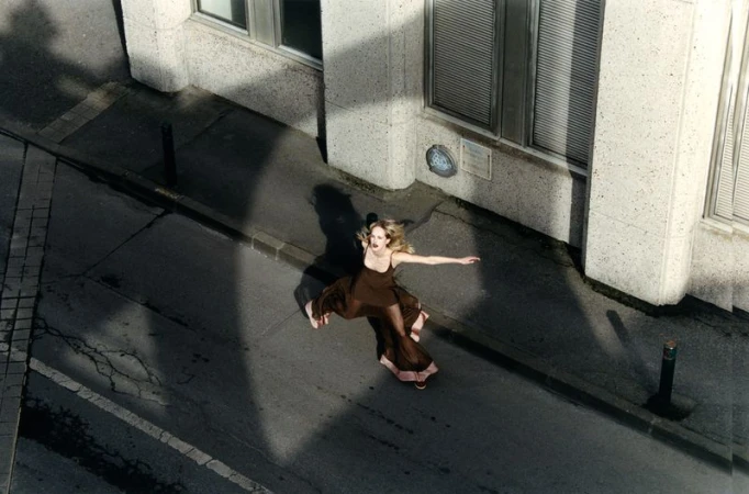 High-angle shot of model Jean Campbell in a brown dress leaping across a city street next to a concrete building.