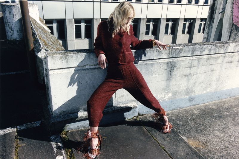 Fashionable woman in burgundy outfit poses on rooftop with building backdrop, showcasing unique footwear and contemporary urban style.