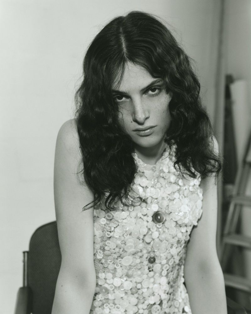 Monochrome portrait of an adult woman with long wavy hair and a sleeveless top with decorative buttons, seated in a chair indoors.