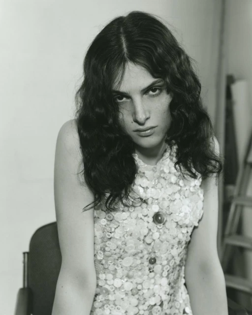 Monochrome portrait of an adult woman with long wavy hair and a sleeveless top with decorative buttons, seated in a chair indoors.