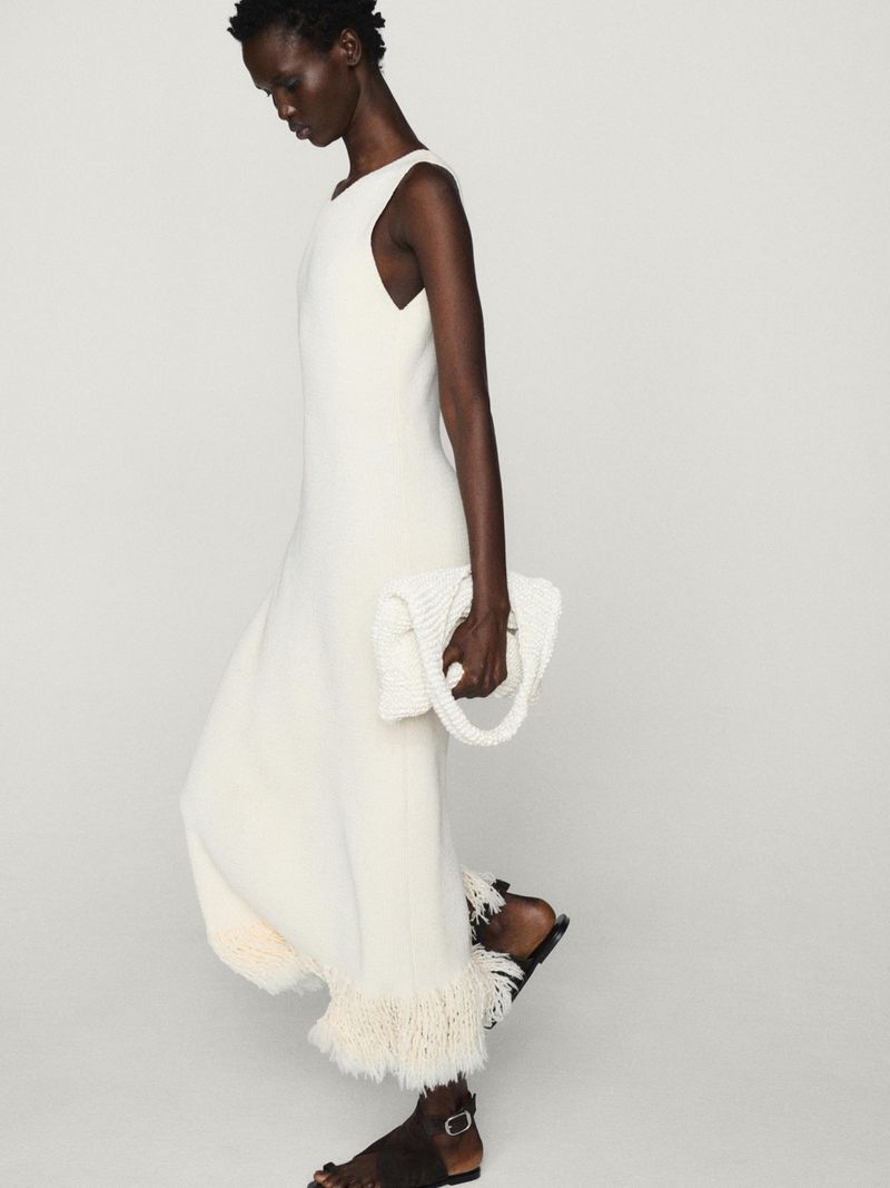 Elegant African American woman in a cream-colored dress and sandals, holding a beaded purse against a neutral studio background.